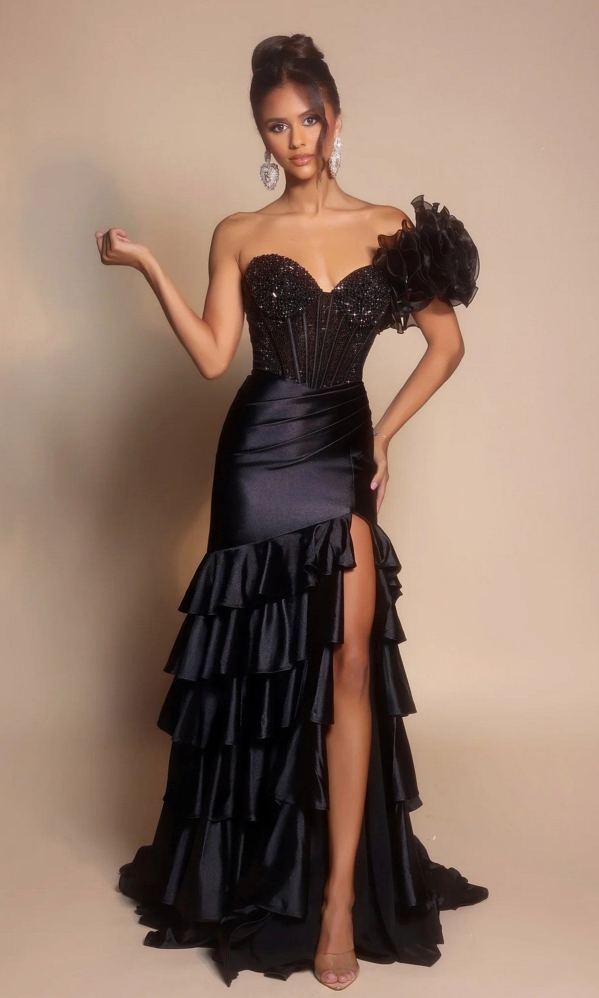 A woman poses in the black Portia and Scarlett PS26141 long formal dress, featuring a beaded corset bodice, strapless mermaid silhouette, satin tiered ruffle skirt with thigh-high slit, and a large fabric flower on one shoulder.